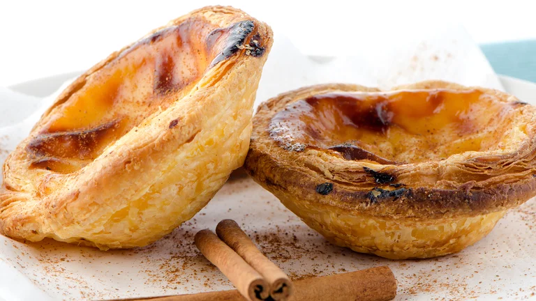 Two Portuguese custard tarts with cinnamon sticks on a white plate.