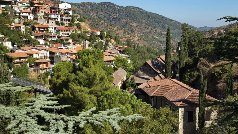 Hillside village with red-roofed houses, green trees, and mountains in the background under a blue sky.