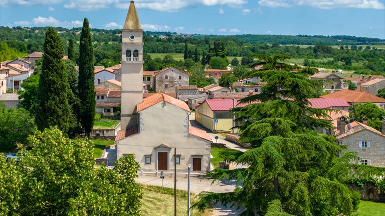 Village with a stone church and bell tower surrounded by trees and countryside under blue sky.