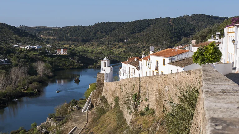 White houses with red roofs line a hill above a river, surrounded by green hills.