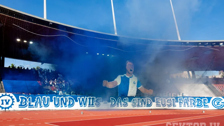Soccer stadium with blue smoke, large fan banner, and cheering crowd in the stands.