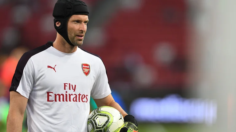 Soccer goalkeeper in Arsenal gear holding a ball, wearing a protective helmet.