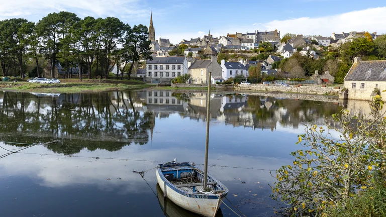 Small boat on calm water with a village and trees reflected in the background.