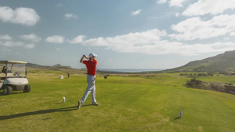 Golfer in red shirt swinging club on green fairway with a golf cart nearby.