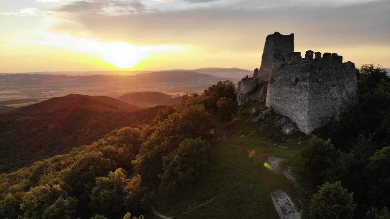 Ruined stone castle atop a forested hill at sunset, with mountains in the background.