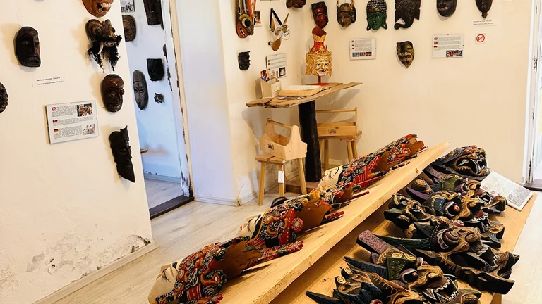 Room filled with colorful masks on walls and tables, wooden floor, and a mannequin at a desk.