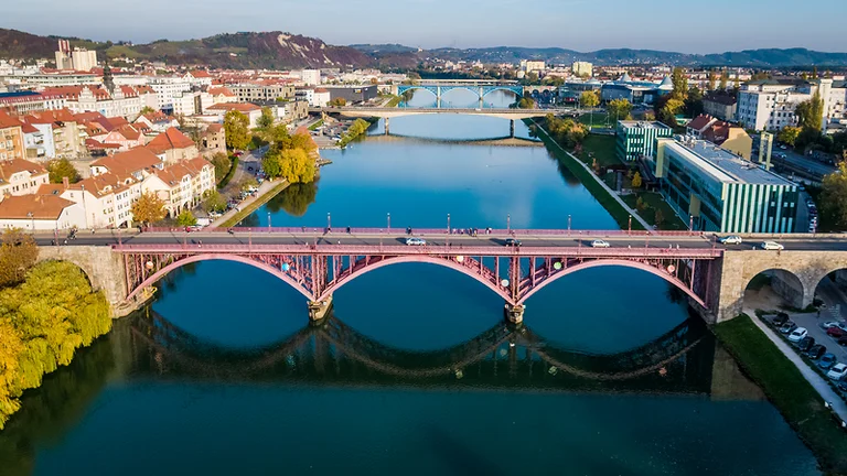 Aerial view of a red arch bridge over a calm river in a city with autumn trees.