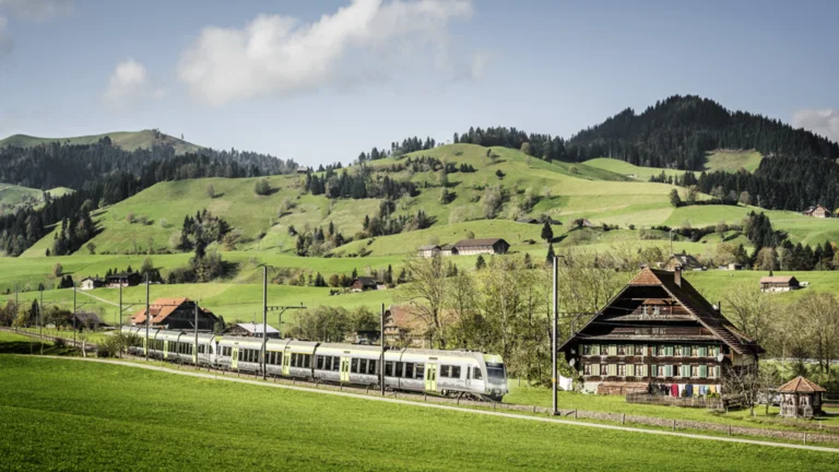 Train passing by traditional Swiss houses with green hills and mountains in the background.