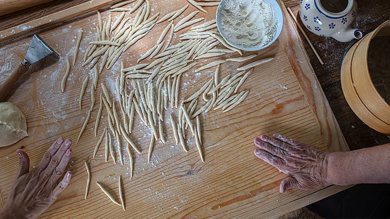 Hands shaping fresh pasta on a floured wooden surface with bowls and dough nearby.