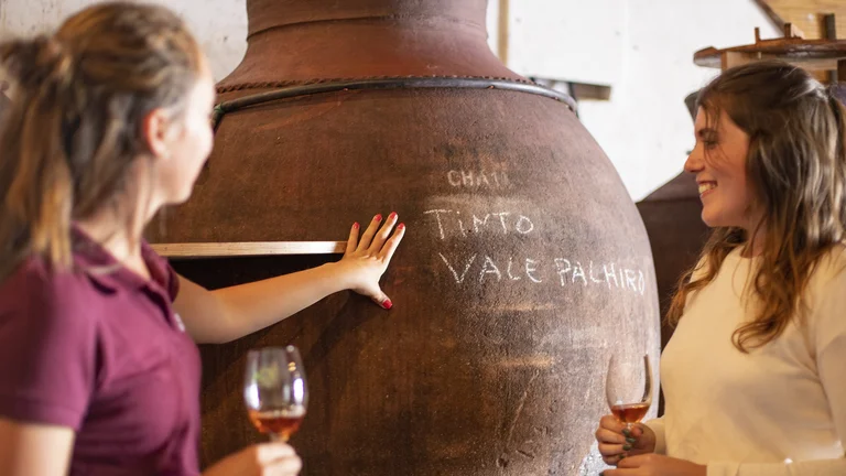 Two women holding wine glasses smile beside a large clay wine vessel with chalk writing.