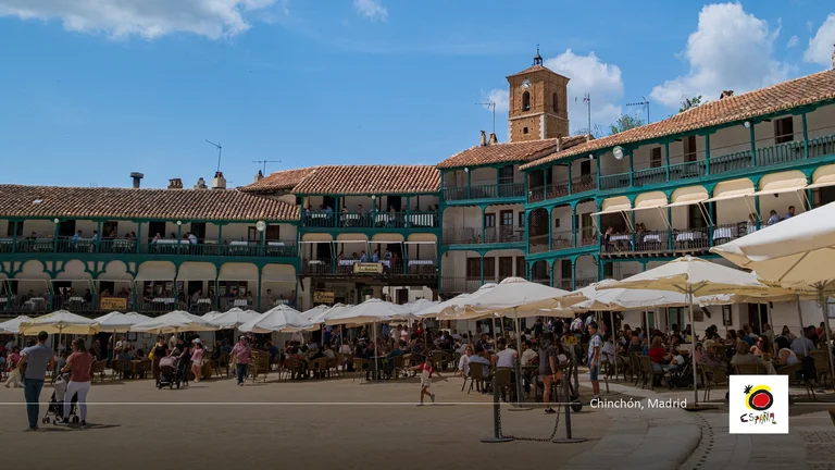 Crowded plaza with umbrellas, people, and colorful historic buildings under a blue sky.