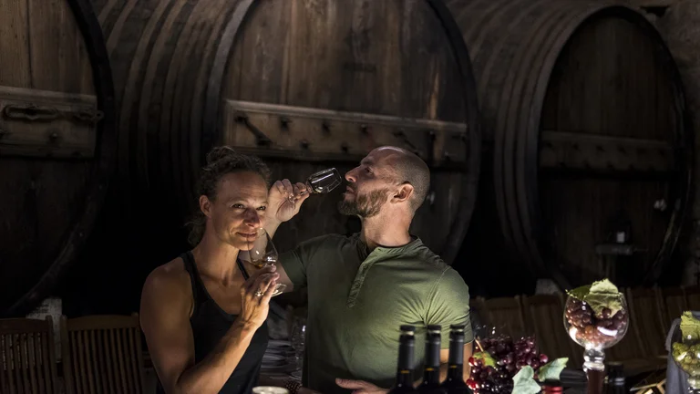 Two people tasting wine in a dimly lit cellar with large wooden barrels.