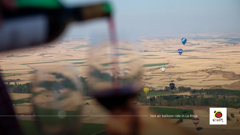 Pouring red wine into a glass with hot air balloons over a vast landscape in the background.