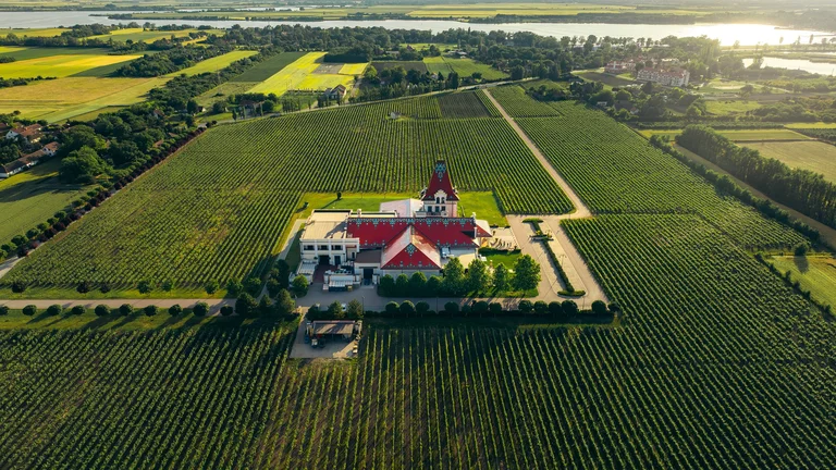 Large building with a red roof surrounded by green farmland, trees, and distant water.