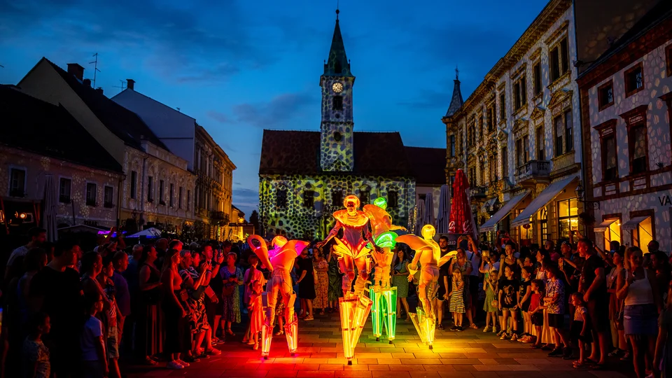 Street performers on stilts lit by colorful lights, surrounded by a crowd at dusk.