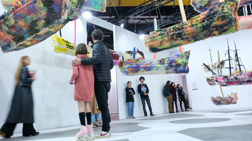 People viewing colorful suspended boat sculptures in a modern art gallery.