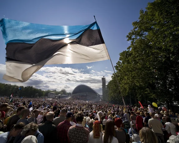 A large Estonian flag waves above a dense crowd of spectators at an outdoor event.