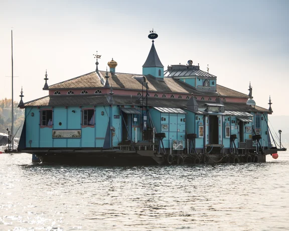 A blue floating house with a teal exterior and decorative roof elements on water.