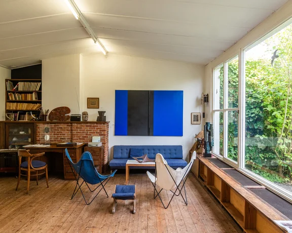 House interior, featuring some blue chairs and sofa, a blue painting, a bookshelf and a desk. A white background and on the right a window.