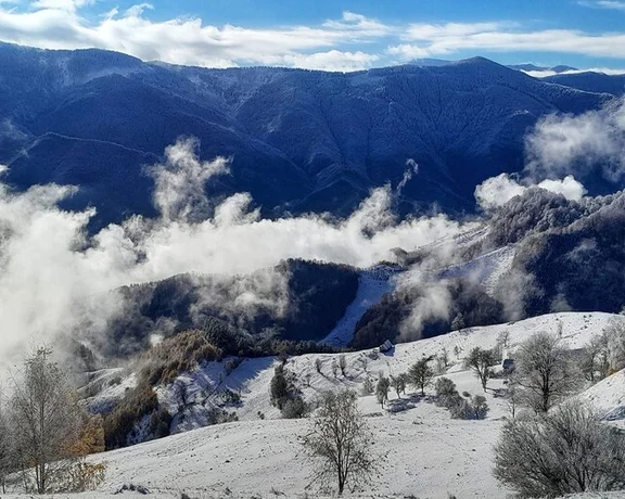 Snow-covered landscape with clouds nestled in mountain valleys under a blue sky.