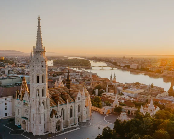 Aerial view of the iconic Matthias Church with its Gothic spire and colorful roof, overlooking the Danube River and the golden cityscape of Budapest at sunrise.