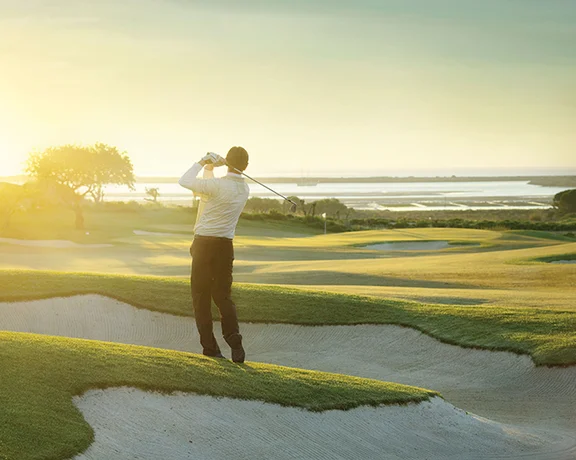 Golfer swinging on a coastal course in Portugal at sunset, with sand bunkers and sea in the background.