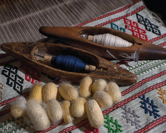 Traditional weaving tools on a patterned fabric, featuring spindles, threads, and natural silkworm cocoons.
