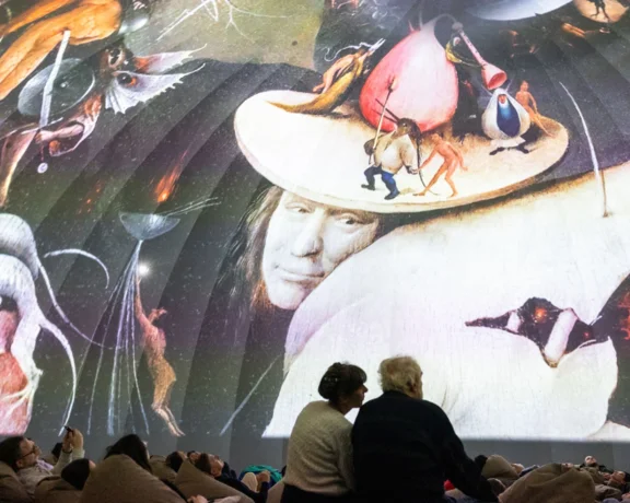 People viewing a large projected artwork with surreal and classical elements in a dim room.