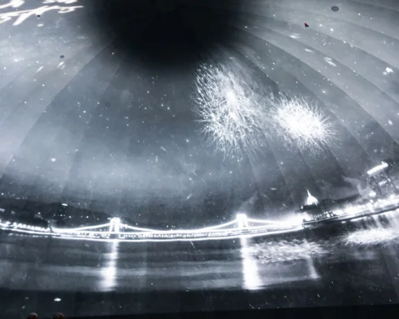 A black and white image showcasing fireworks above a lit bridge, reflected on a surface, with spectators silhouetted below.