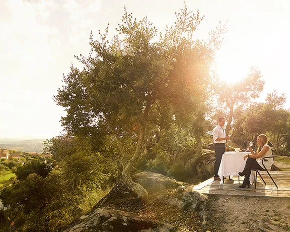 Couple enjoying an outdoor meal near a wooden cabin overlooking a scenic village at sunset.