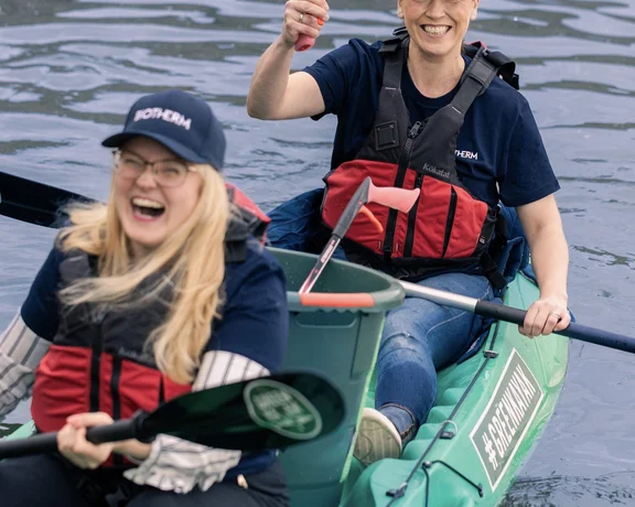 Two women kayaking with a green kayak, smiling and collecting waste from the water in a shared kayak.