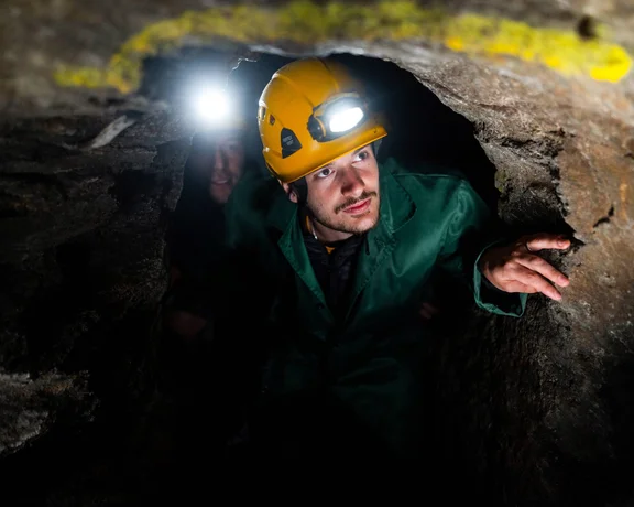 Person in a green jacket and helmet with a headlamp emerging from a dark cave.