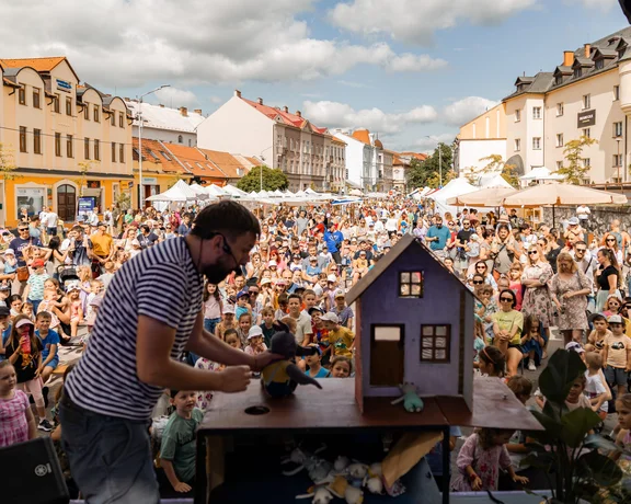 A man playing with a puppet, standing on a stage in front of a crowd in a colorful square.