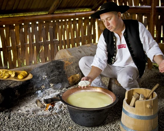 A man demonstrates traditional cooking methods inside a rustic wooden hut, with a pot over a fire and various tools and food nearby.