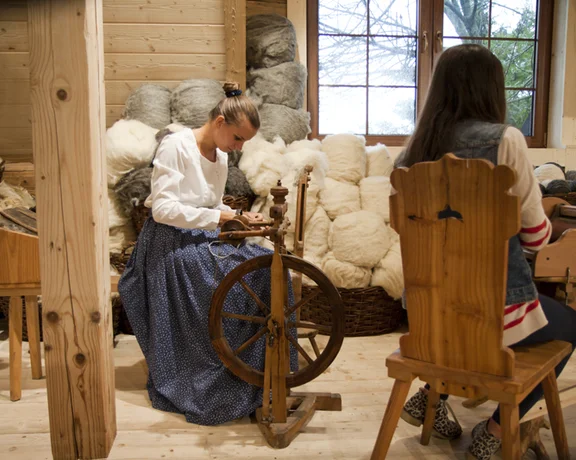 Two women work in a rustic workshop, one spinning yarn on a wheel, surrounded by baskets of wool. Natural light pours in from the window.