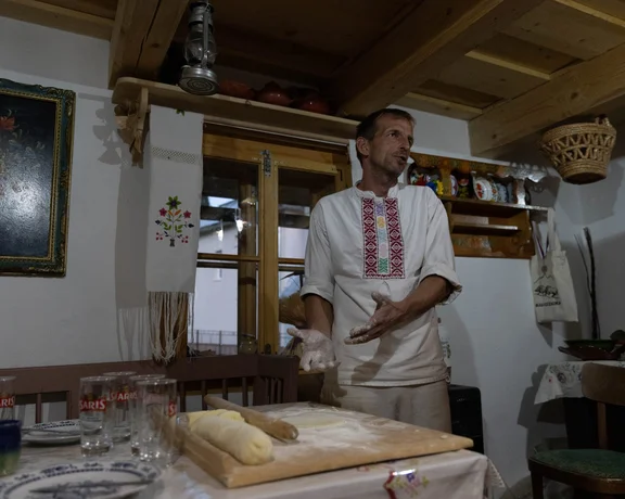 A person in traditional attire stands by a wooden table with dough, surrounded by rustic kitchen decor and glassware.