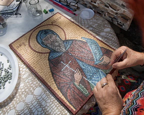 A person carefully works on a mosaic of a saint, surrounded by mosaic tiles and tools on a table.