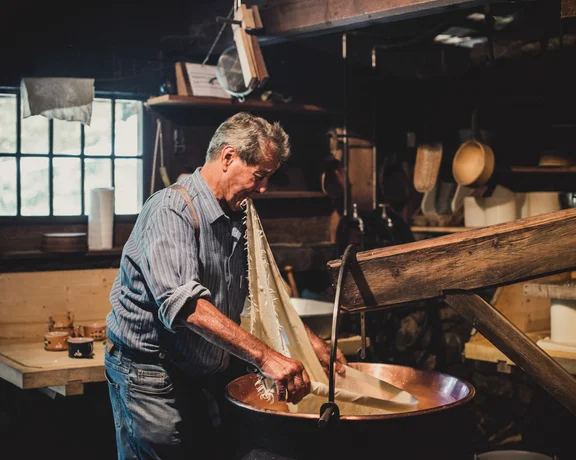 A man in a striped shirt works with fabric in a rustic workshop, surrounded by wooden tools and shelves filled with pottery and supplies.
