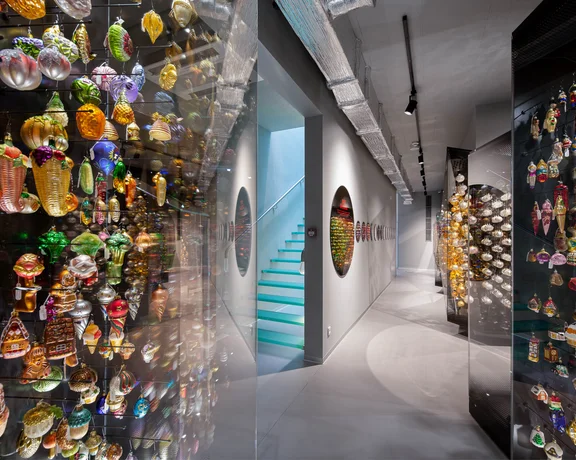 A colorful display of vintage glass ornaments hangs on glass walls in a modern hallway, leading to turquoise stairs.
