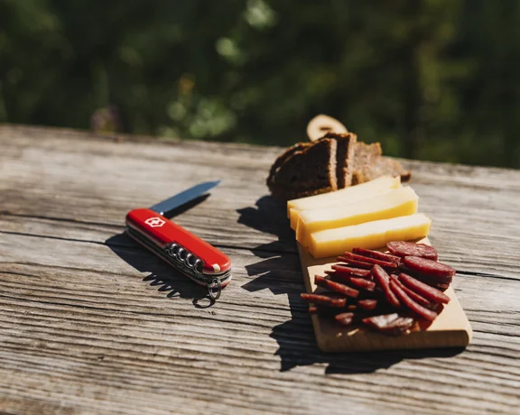A red Swiss knife beside a wooden board with slices of cheese, cured meat, and dark bread on a rustic wooden table in nature.