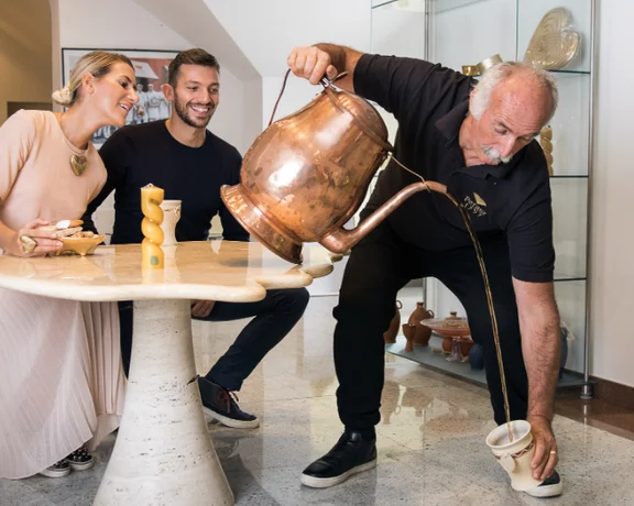 A man pours liquid from a large copper pot into a small mug at a table with two seated people, showcasing a cultural experience.