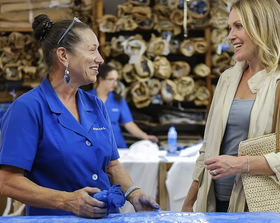A woman in a blue shirt demonstrates fabric dyeing techniques to a visitor, surrounded by rolls of fabric in a workshop.