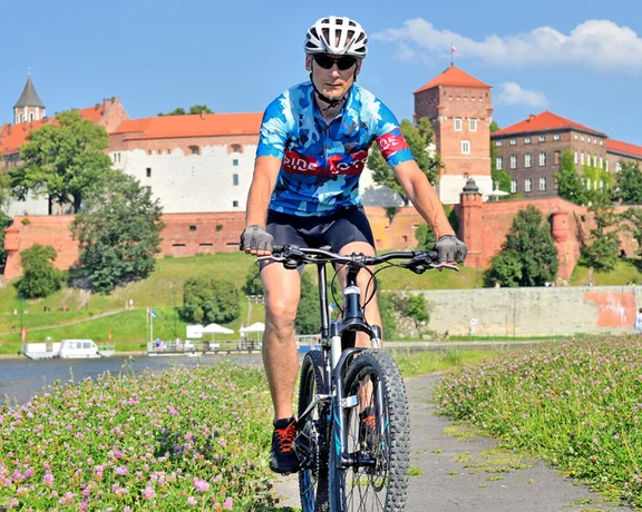 A cyclist on a path lined with flowers rides near a historic castle under a clear blue sky in Kraków.