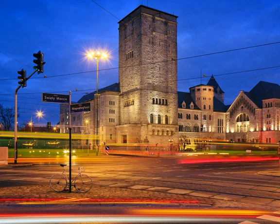 Illuminated historic building in Poznań at twilight with traffic lights, a bicycle, and motion-blurred trams in the foreground.