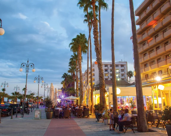 A lively promenade at dusk, lined with palm trees, cafes, and cars, illuminated by street lamps under a cloudy sky in Larnaka.