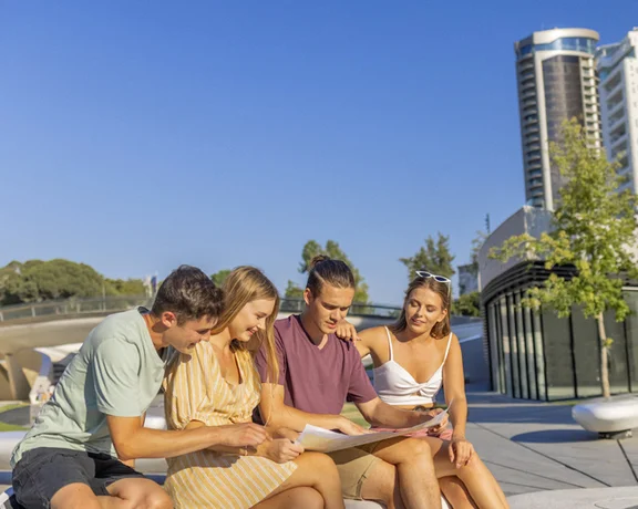 Four friends gather in Eleftheria Square in Nicosia, studying a map with modern buildings and greenery in the background under a clear blue sky.