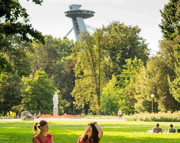 Two young women sitting on a blanket in a green park enjoying a sunny day, with Bratislava's UFO Tower visible in the background.