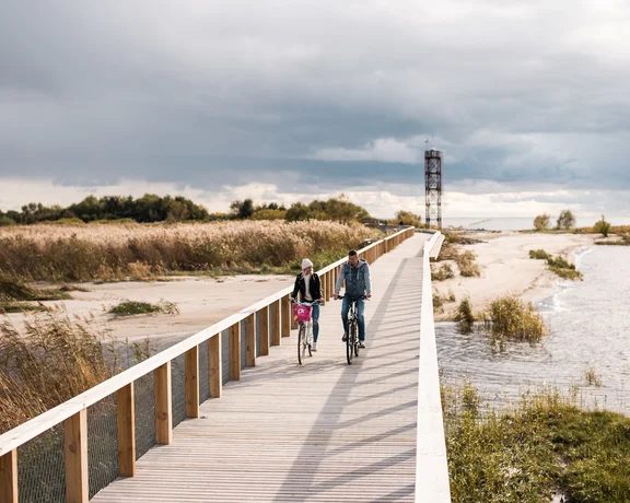 A couple rides bicycles along a wooden boardwalk beside a serene river and sandy landscape, under a cloudy sky.