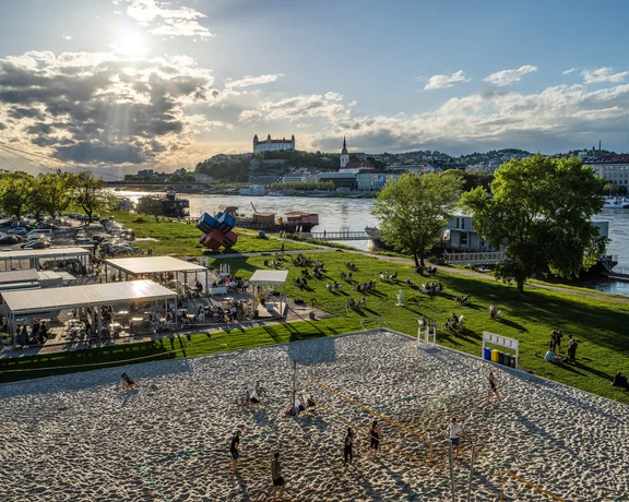 A lively riverside scene with people enjoying beach volleyball and picnicking, with a view of a bridge and Bratislava Castle in the background.