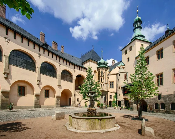 A historic courtyard featuring elegant architecture, a stone fountain, and trees under a bright blue sky with fluffy clouds.